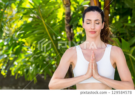 Woman meditating with hands pressed in garden among tropical leaves wearing white sports top Woman meditating with hands pressed in garden among tropical leaves wearing white sports top 129584578