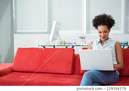African American woman typing on laptop on red sofa in office lounge with monitor, copy space 129584579