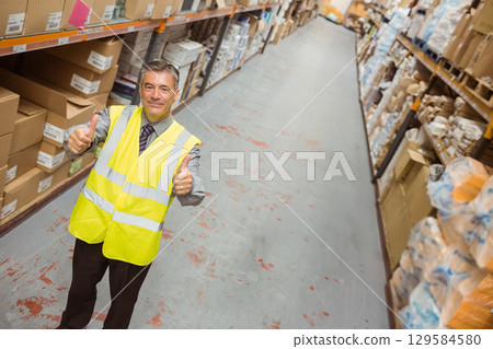 Male warehouse supervisor giving thumbs up wearing safety vest in aisle among racks, copy space 129584580
