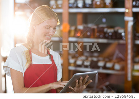 Female employee wearing red apron updating stock on tablet in aisle with shelves, jars, copy space 129584585