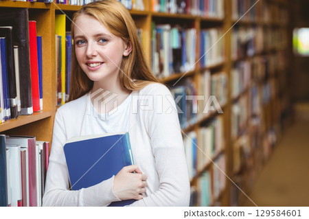 Teenage female student holding blue hardcover book in library aisle with wooden shelves, copy space Teenage female student holding blue hardcover book in library aisle with wooden shelves, copy space 129584601