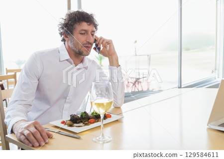 Man talking on smartphone at restaurant table with laptop, meat plate and wine glass, copy space Man talking on smartphone at restaurant table with laptop, meat plate and wine glass, copy space 129584611