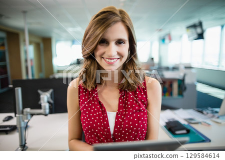Woman smiling while typing on laptop at modern office desk with monitor arm wearing red blouse 129584614