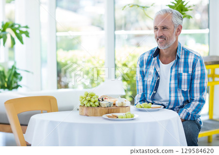 Senior man sitting at table near windows, admiring cheeseboard with grapes, crackers, copy space 129584620