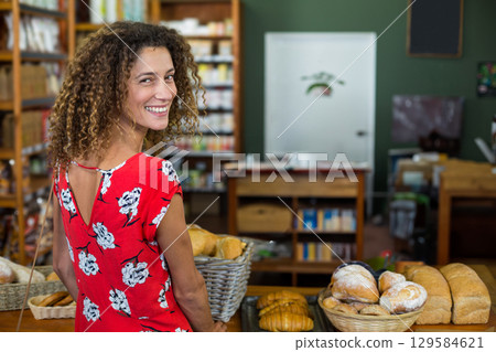 Smiling adult woman standing behind bakery counter displaying wicker baskets of loaves, copy space 129584621