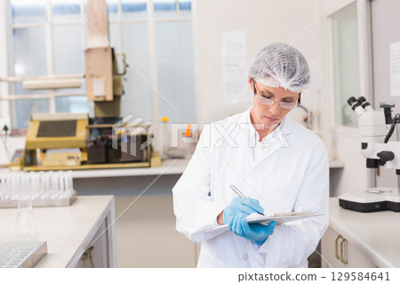 Female scientist wearing lab coat writing on clipboard at bench with test tube rack, copy space Female scientist wearing lab coat writing on clipboard at bench with test tube rack, copy space 129584641