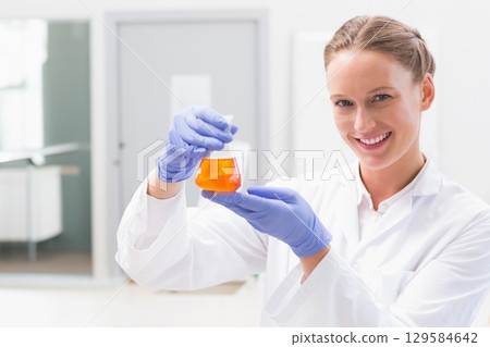 Female scientist wearing coat, gloves, holding orange liquid in Erlenmeyer flask in lab, copy space 129584642