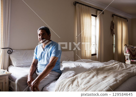 Senior man sitting on bed edge in bedroom holding water glass near nightstand, copy space 129584645
