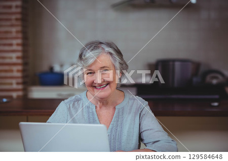 Senior woman typing on laptop at kitchen counter with blue pot and tiled backsplash 129584648