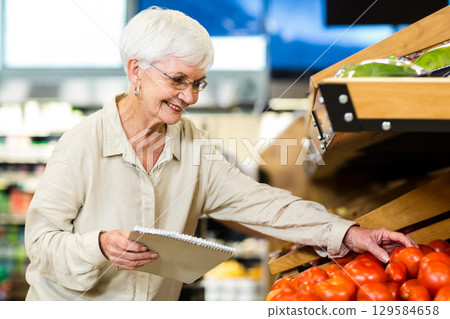 Senior female shopper picking ripe red tomato in supermarket produce bin holding spiral notepad 129584658