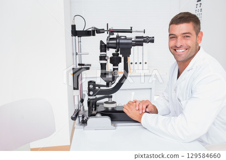 Male optometrist wearing lab coat sitting beside slit lamp biomicroscope at clinic desk, copy space 129584660