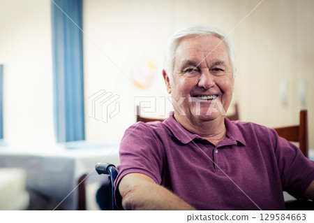 Senior man smiling while sitting in wheelchair at dining room with white tablecloth, blue curtains Senior man smiling while sitting in wheelchair at dining room with white tablecloth, blue curtains 129584663