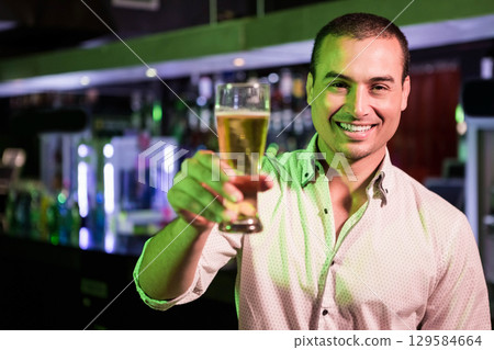 Man standing at bar holding beer glass by counter with liquor bottles, LED lights, copy space 129584664