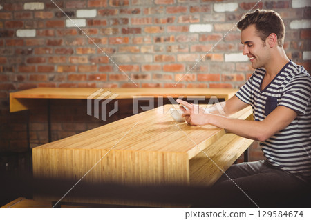 Middle-aged man sitting at cafe table holding mug and touching smartphone by brick wall, copy space 129584674