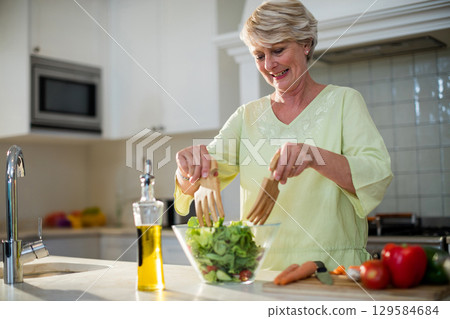 Senior woman tossing green salad in home kitchen using glass bowl and wooden servers, copy space Senior woman tossing green salad in home kitchen using glass bowl and wooden servers, copy space 129584684