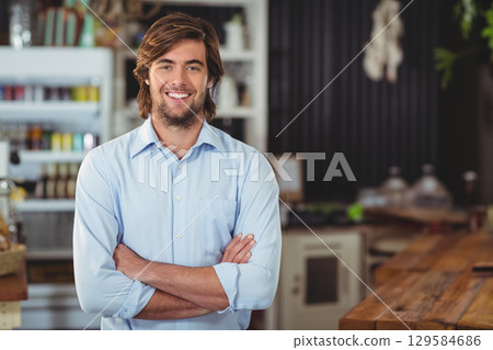 Bearded man standing with arms crossed in cafe with wooden service counter and glass cake domes 129584686