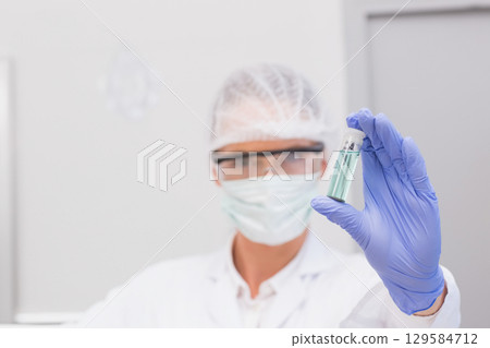 Gloved hand holding small clear vial containing green liquid in sterile lab, near grey cabinet 129584712