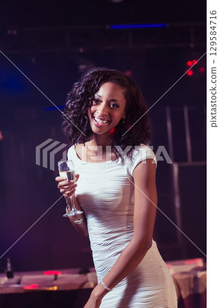 African American woman wearing white dress raising champagne flute in club with tables and lighting 129584716