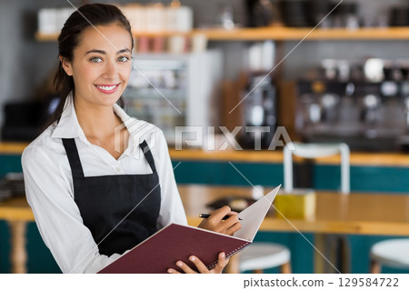 Female barista holding pen over notebook behind wooden counter at coffee shop, copy space 129584722