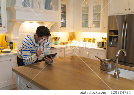 Middle-aged man leaning on kitchen island using tablet while stirring saucepan with wooden spoon 129584723