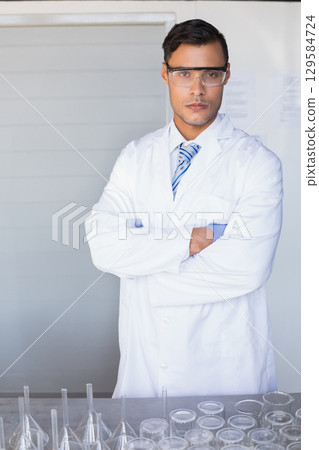 Male scientist wearing lab coat standing with crossed arms behind bench displaying glassware in lab 129584724