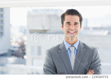 Male professional standing arms crossed in grey suit and patterned tie by large window, copy space Male professional standing arms crossed in grey suit and patterned tie by large window, copy space 129584733