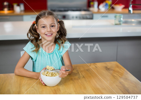 School-age girl holding metal spoon over white bowl and eating cereal at modern kitchen table School-age girl holding metal spoon over white bowl and eating cereal at modern kitchen table 129584737