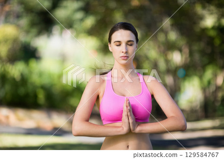 Woman wearing pink sports bra standing palms pressed together meditating in park under sunlight 129584767
