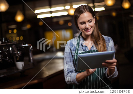 Woman wearing striped apron tapping tablet behind counter at cafe with espresso machine, copy space 129584768