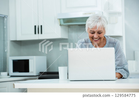 Senior woman wearing grey blouse using laptop at kitchen island with microwave and ceramic mug Senior woman wearing grey blouse using laptop at kitchen island with microwave and ceramic mug 129584775