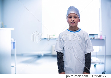Boy standing in treatment room wearing surgical cap and gown by window and cart, copy space 129584776