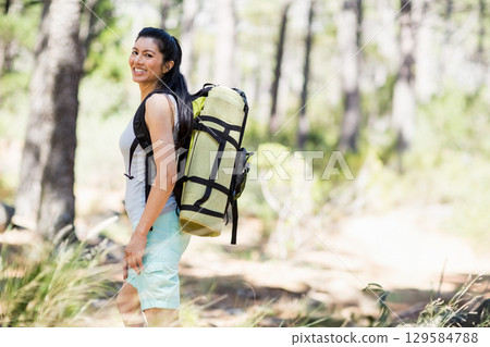 Asian woman hiker looking over shoulder toward camera on sunlit pine dirt trail with green backpack 129584788