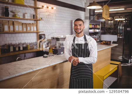 Male barista standing behind marble counter at cafe displaying coffee bags and pastries, copy space 129584796