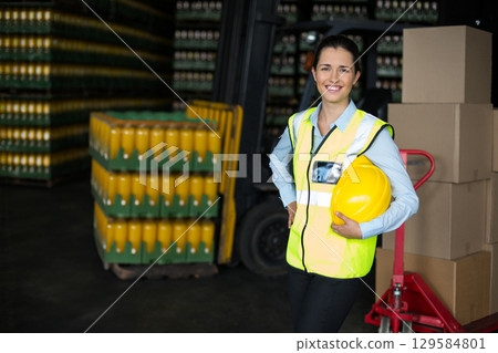 Female warehouse worker wearing safety vest holding hard hat amid beverage crates, copy space Female warehouse worker wearing safety vest holding hard hat amid beverage crates, copy space 129584801