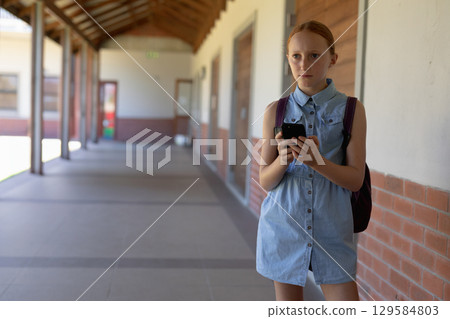 Girl student standing in school corridor holding smartphone and backpack, copy space Girl student standing in school corridor holding smartphone and backpack, copy space 129584803