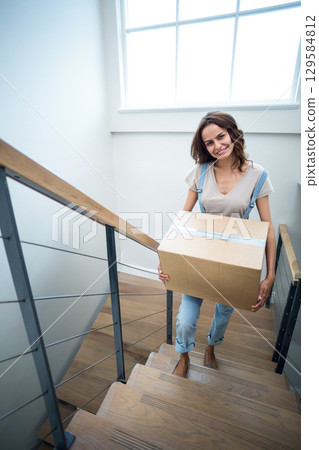 Mid adult woman in overalls carrying taped box up home staircase with metal railing under window Mid adult woman in overalls carrying taped box up home staircase with metal railing under window 129584812