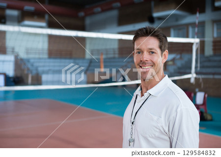 Male coach standing near volleyball net on gymnasium court holding whistle on lanyard, copy space 129584832