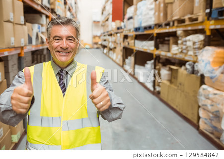 Male warehouse supervisor smiling wearing safety vest and giving thumbs up among shelving and boxes 129584842
