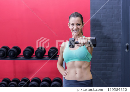 Fit woman in sportswear smiling while lifting dumbbell in weight room with rack of black dumbbells 129584850