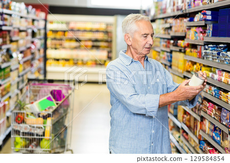 Senior man examining food container label while standing in supermarket aisle with shopping cart Senior man examining food container label while standing in supermarket aisle with shopping cart 129584854