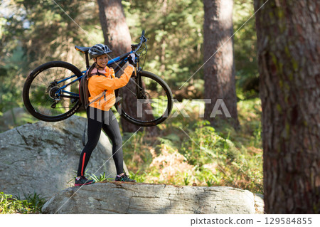 Woman carrying mountain bike on shoulder, navigating forest trail wearing orange jacket and helmet Woman carrying mountain bike on shoulder, navigating forest trail wearing orange jacket and helmet 129584855