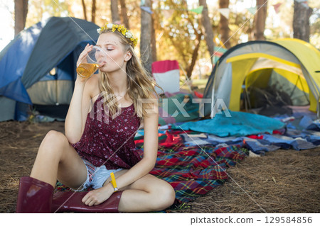 Woman sitting on plaid blanket at forest camp wearing yellow floral crown holding plastic cup 129584856