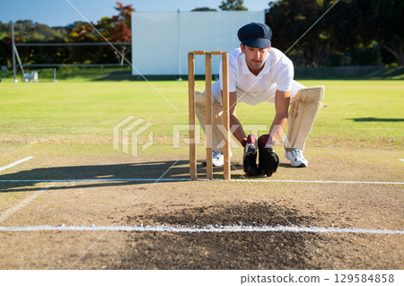 Male wicket keeper wearing white kit crouching behind stumps on pitch gloves pads with sight screen Male wicket keeper wearing white kit crouching behind stumps on pitch gloves pads with sight screen 129584858