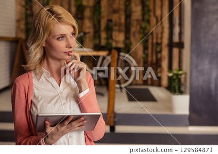 woman wearing blouse standing in modern office holding tablet near bicycle gazing right, copy space 129584871