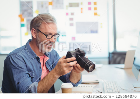 Middle-aged man in denim shirt inspecting DSLR camera at office desk with coffee cup and notebook 129584876