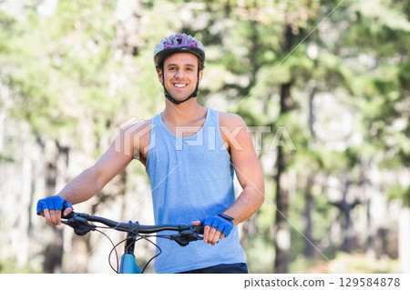 Male cyclist standing smiling on sunlit forest trail holding bike handlebars wearing helmet Male cyclist standing smiling on sunlit forest trail holding bike handlebars wearing helmet 129584878