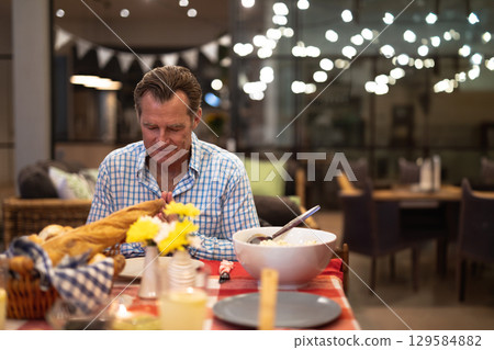 Senior man sitting at cafe dining table praying before meal with wicker bread basket 129584882