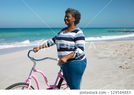 Senior African American woman standing on sandy beach holding pink cruiser bicycle, copy space Senior African American woman standing on sandy beach holding pink cruiser bicycle, copy space 129584883