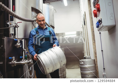 Male brewery worker wearing overalls lifting keg near stainless steel tanks and control panel 129584884
