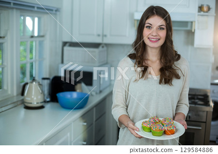 Woman standing in kitchen holding plate of frosted cupcakes with sprinkles by kettle Woman standing in kitchen holding plate of frosted cupcakes with sprinkles by kettle 129584886